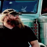 A truck driver with beard and hat standing in front of his vehicle.