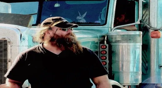 A truck driver with beard and hat standing in front of his vehicle.