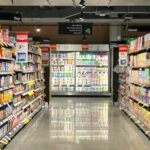 Grocery store aisle with fully stocked shelves of cereal, coffee, and tea, leading back to refrigerated dairy cases under bright overhead lighting.