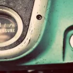 Close-up of a vintage truck dashboard, an analog oil pressure gauge and ignition key on a weathered turquoise panel.